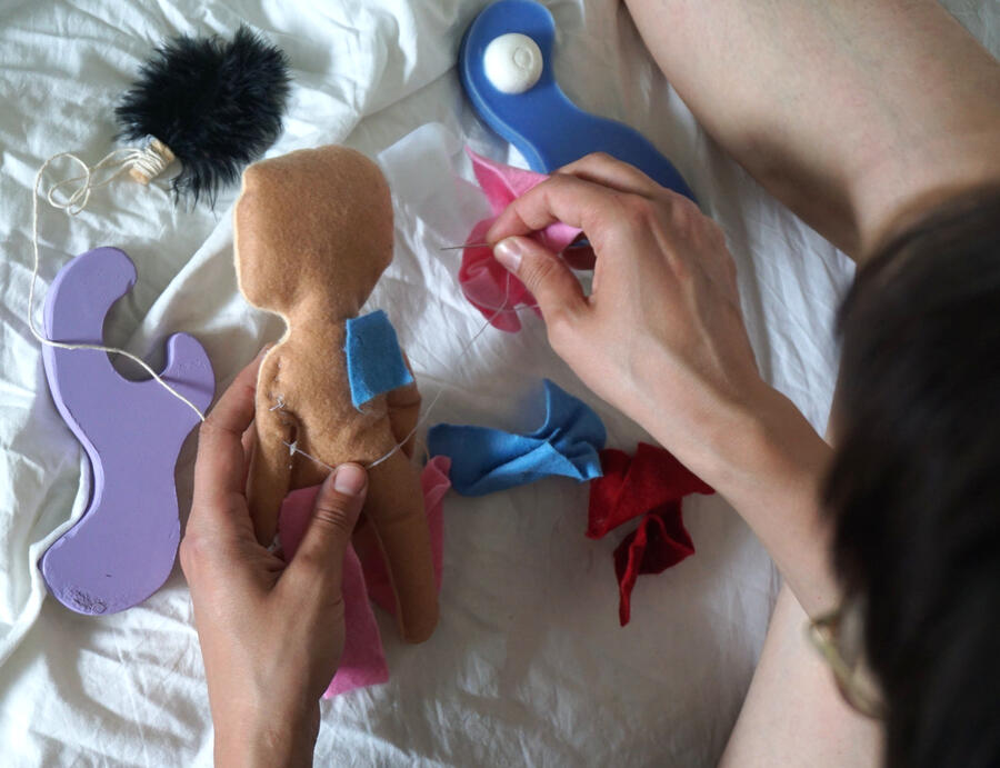 image taken from behind a person who is holding a doll made of felt in their hand. with the other hand they are stitching a piece of blue fabric on the doll. the person is sitting on a white bed sheet and they have multiple objects in front of them.